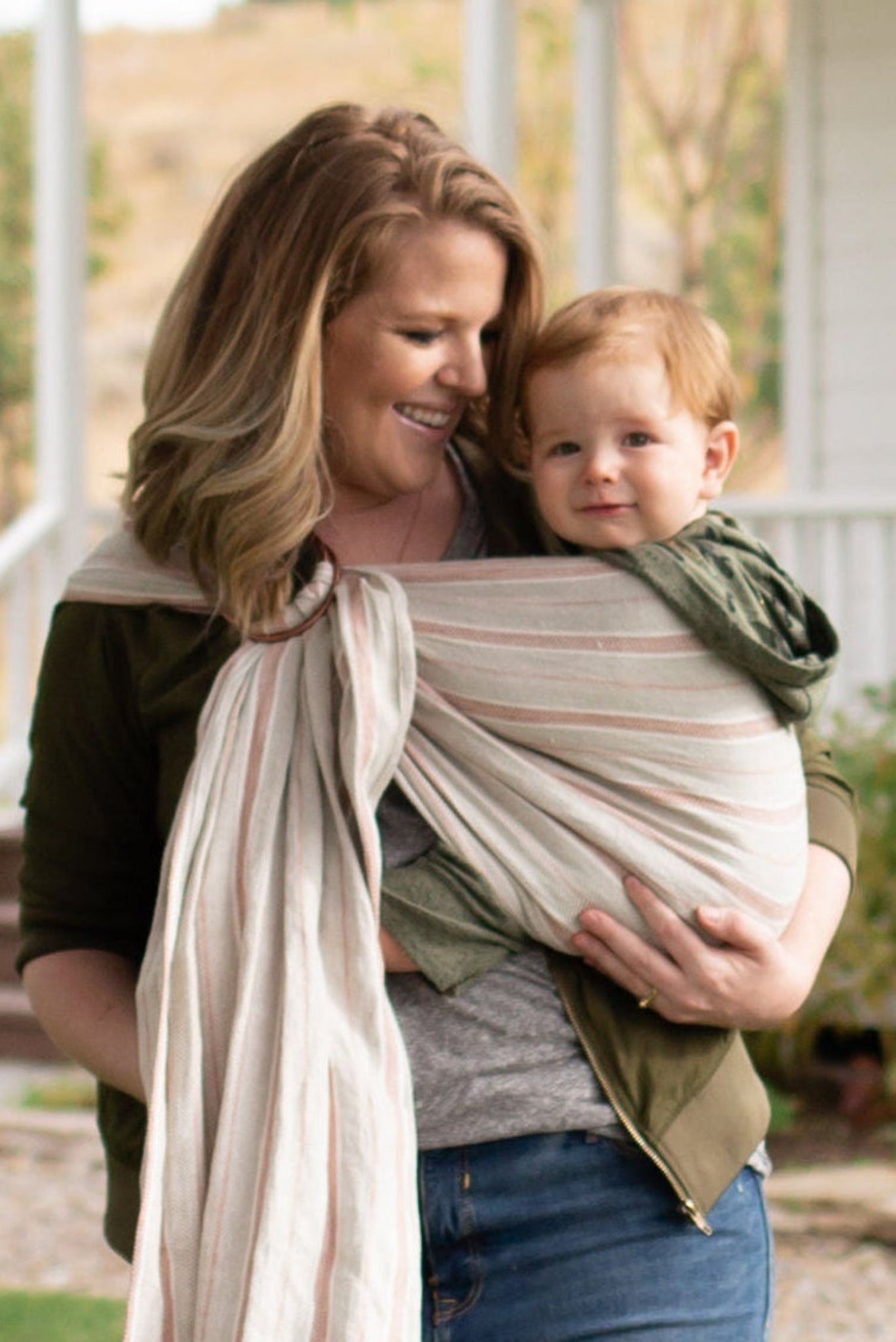 Woman walking outdoors with a child in a sling, house in the background