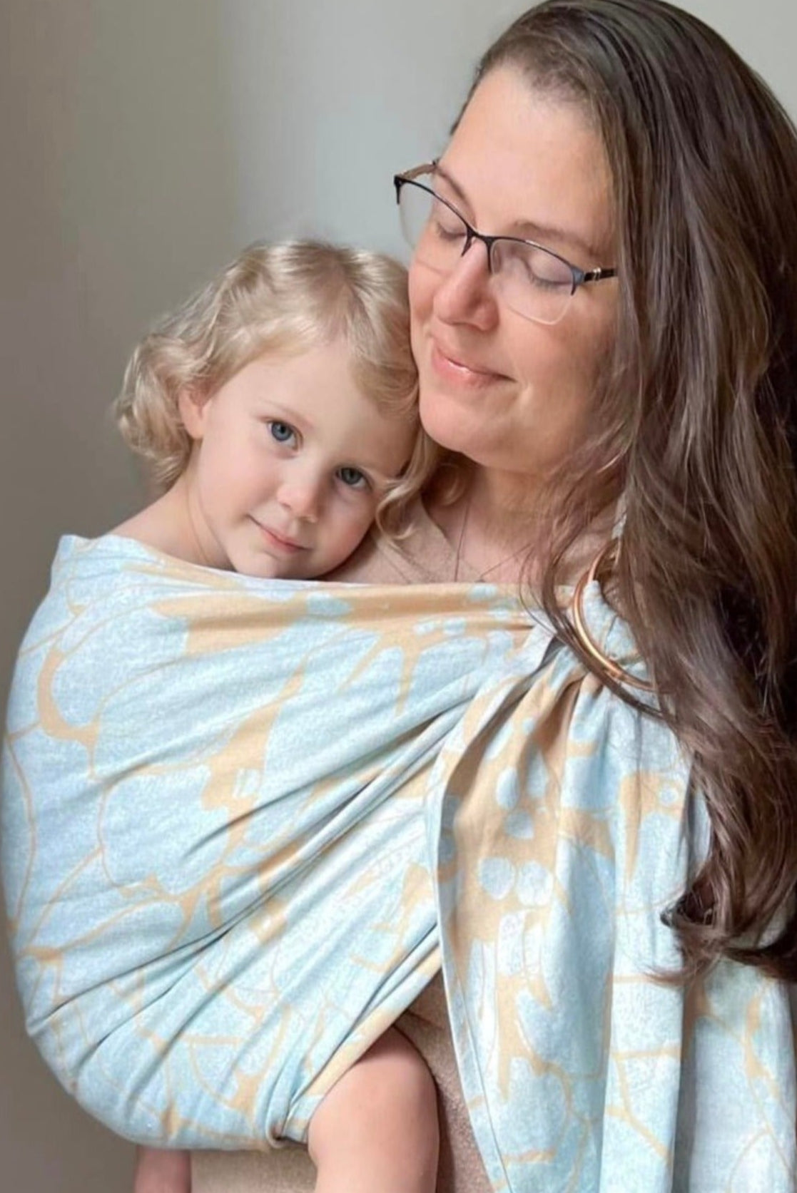 A person is wearing a light blue and gold patterned ring sling baby carrier with a baby inside.
