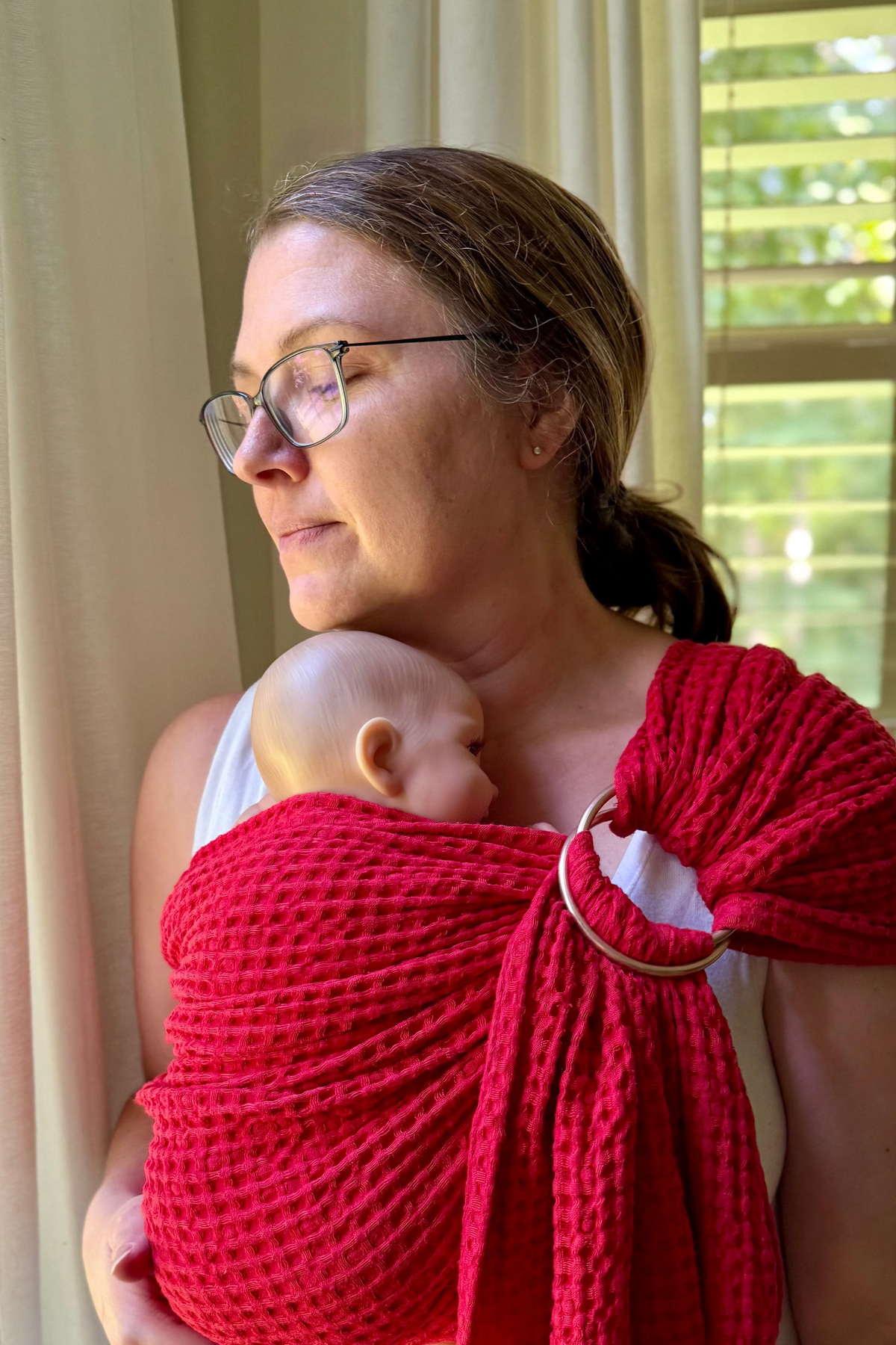 Woman holding a baby wrapped in a red sling in a room with curtains and a window.
