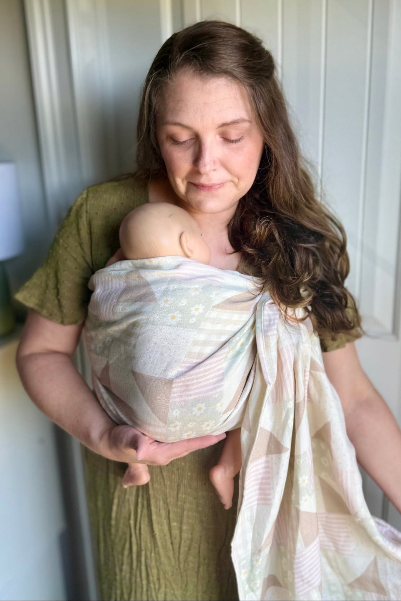 A woman wearing an olive green dress holds a baby wrapped in a light beige and white patterned baby wrap or shawl against a white paneled door.