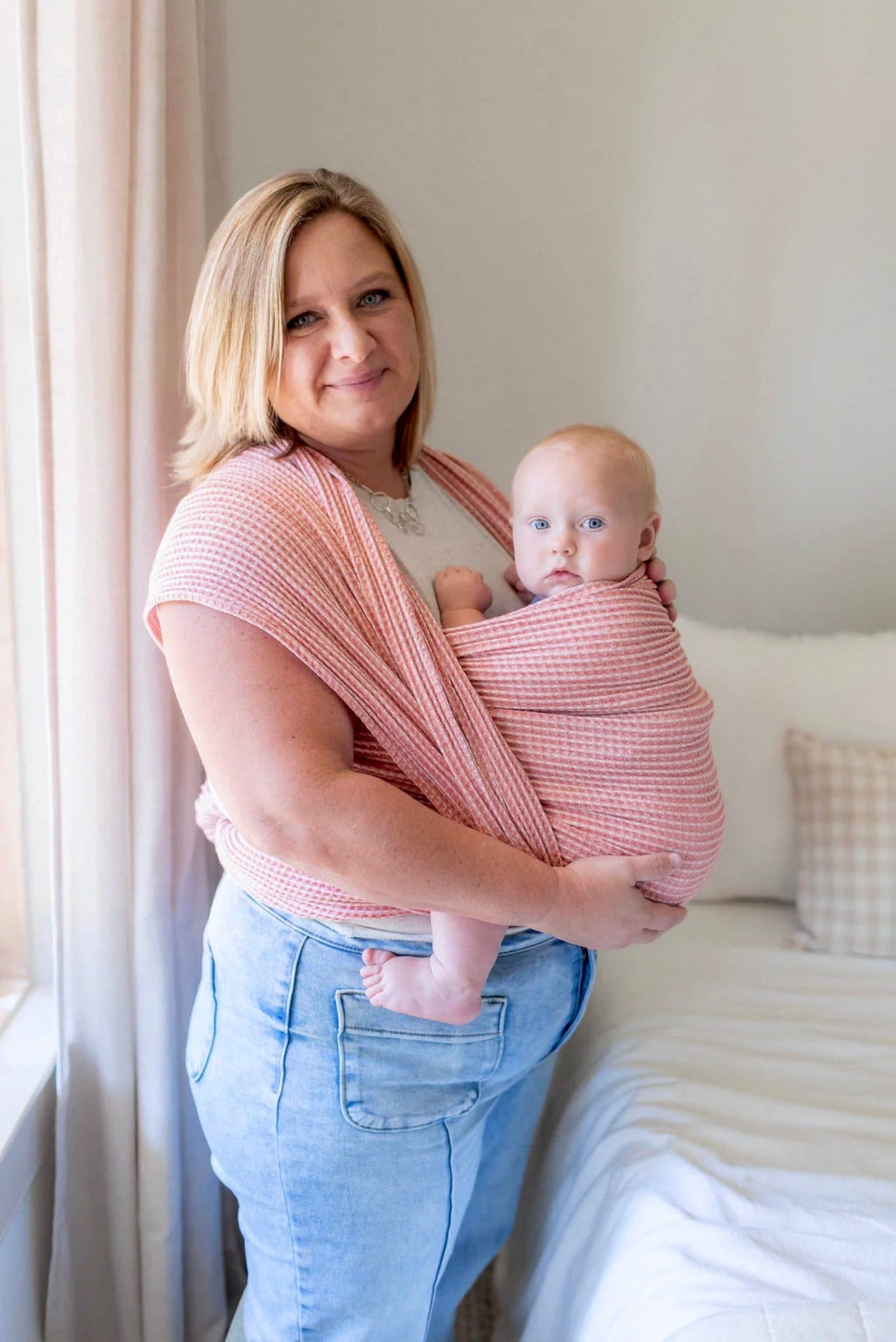 Woman holding a baby in a pink woven wrap in a bedroom setting.