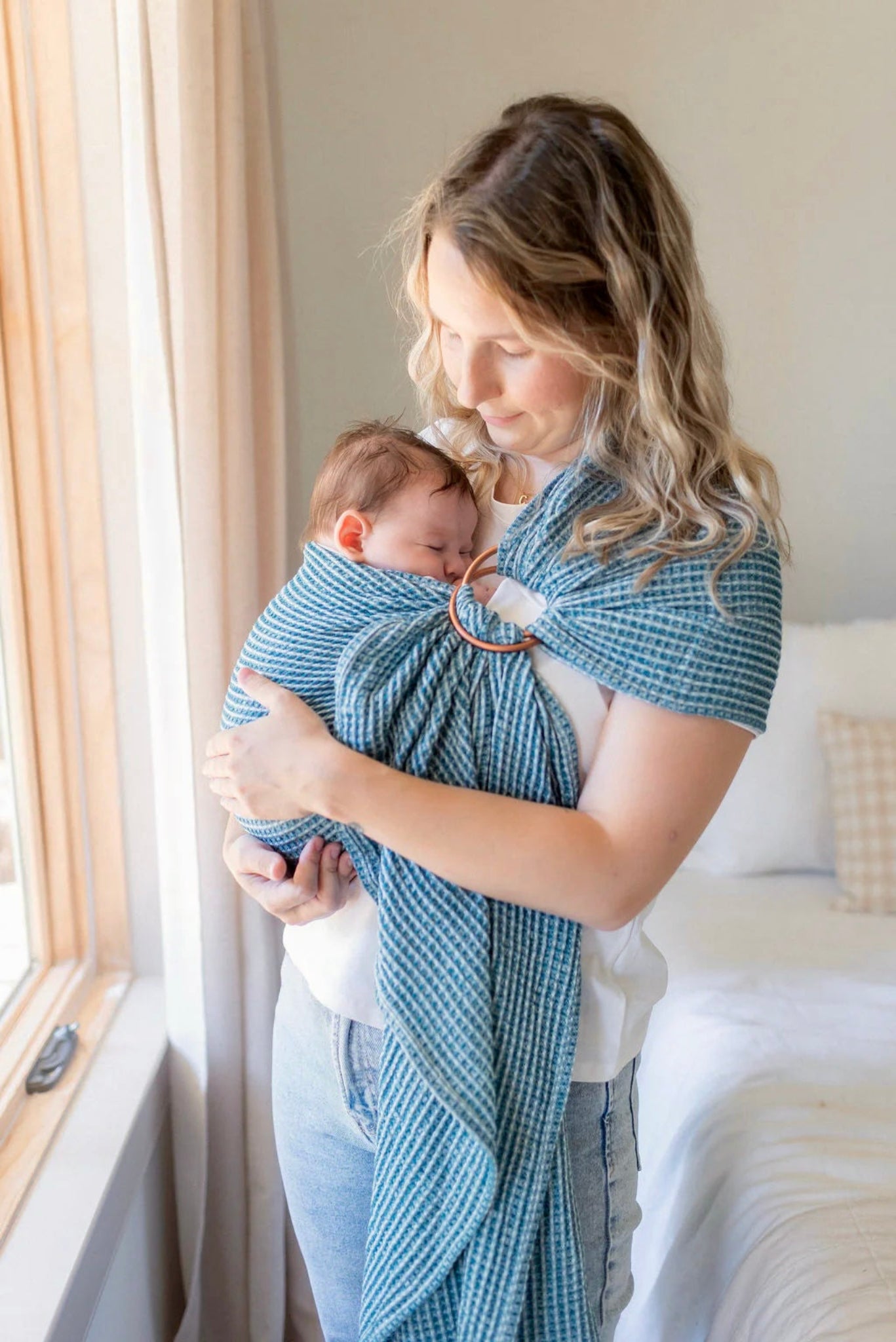 Woman holding a baby wrapped in a teal blue woven wrap in a softly lit room.