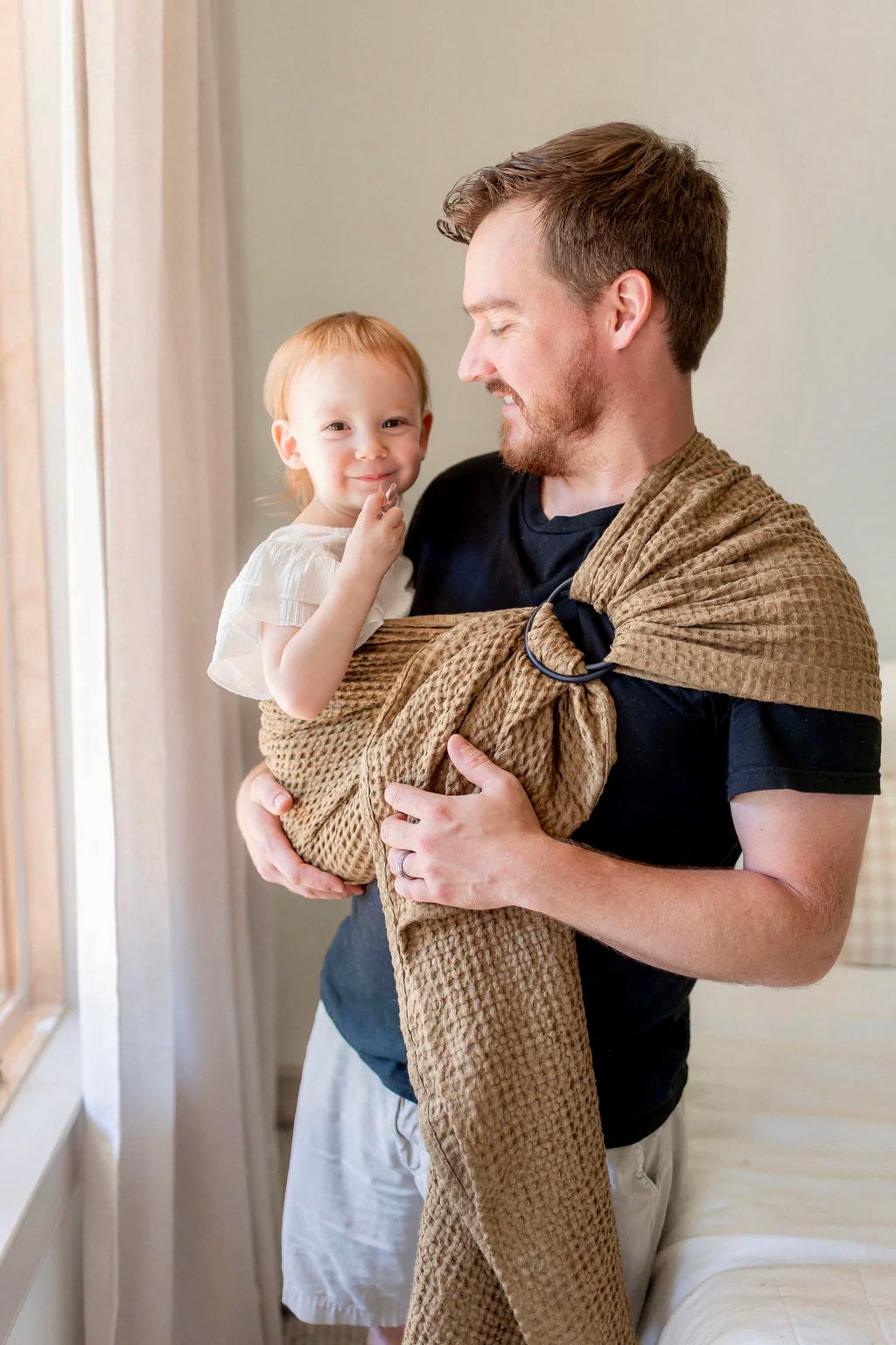 Man holding a child wrapped in a brown woven wrap against a neutral background