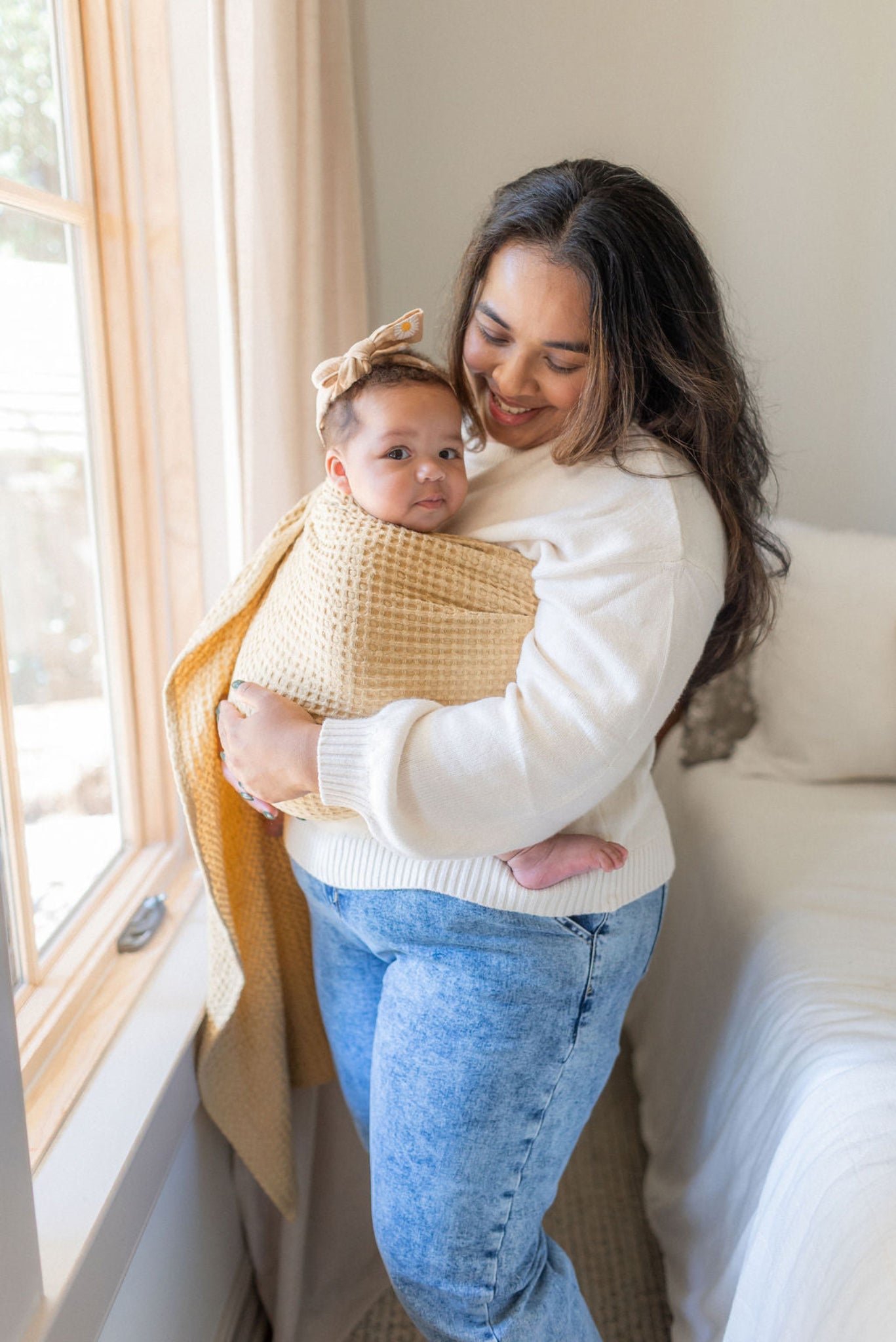 Woman holding a baby in a cozy room with a window and couch.
