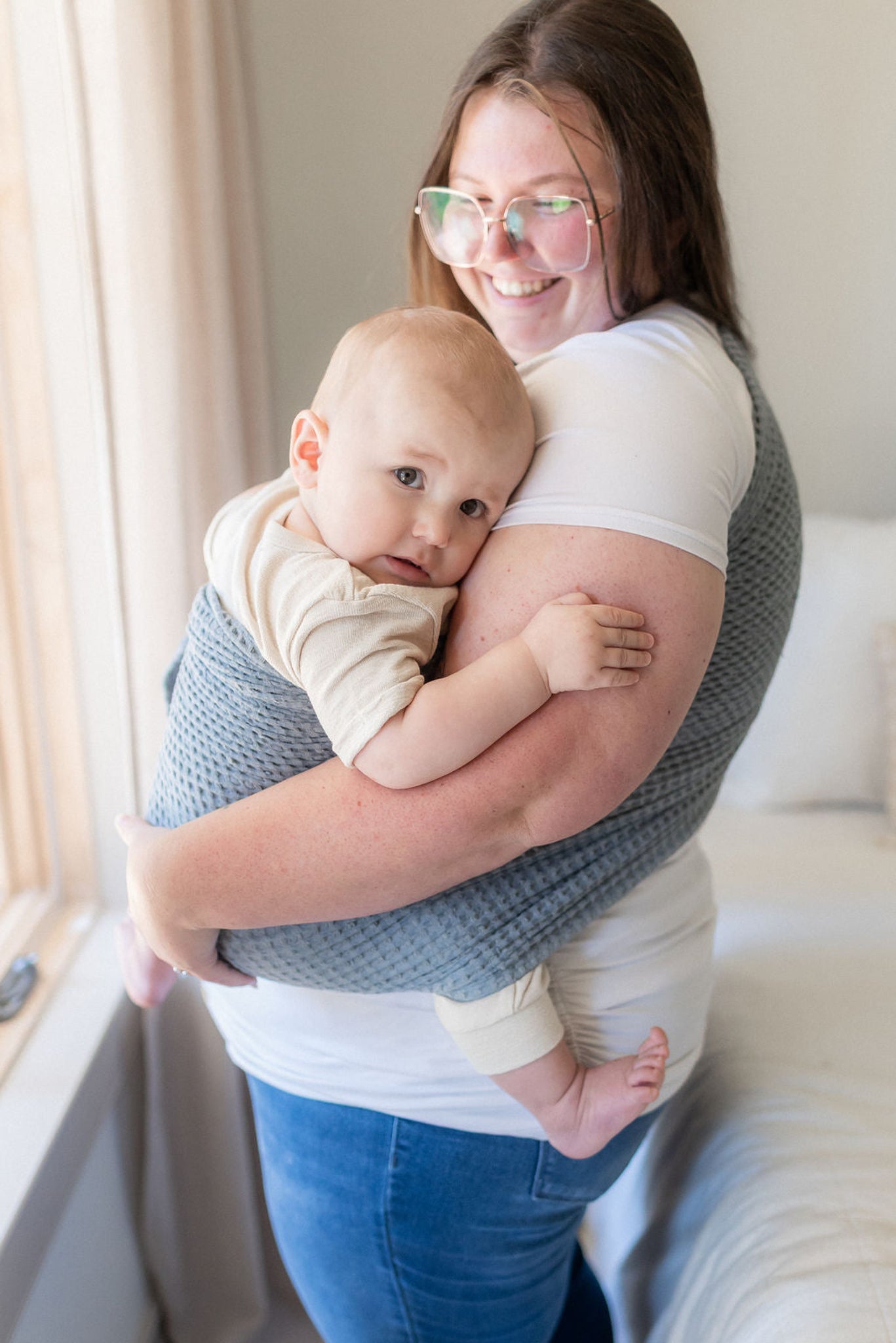 Woman holding a baby wrapped in a gray wrap, smiling in a bright room.