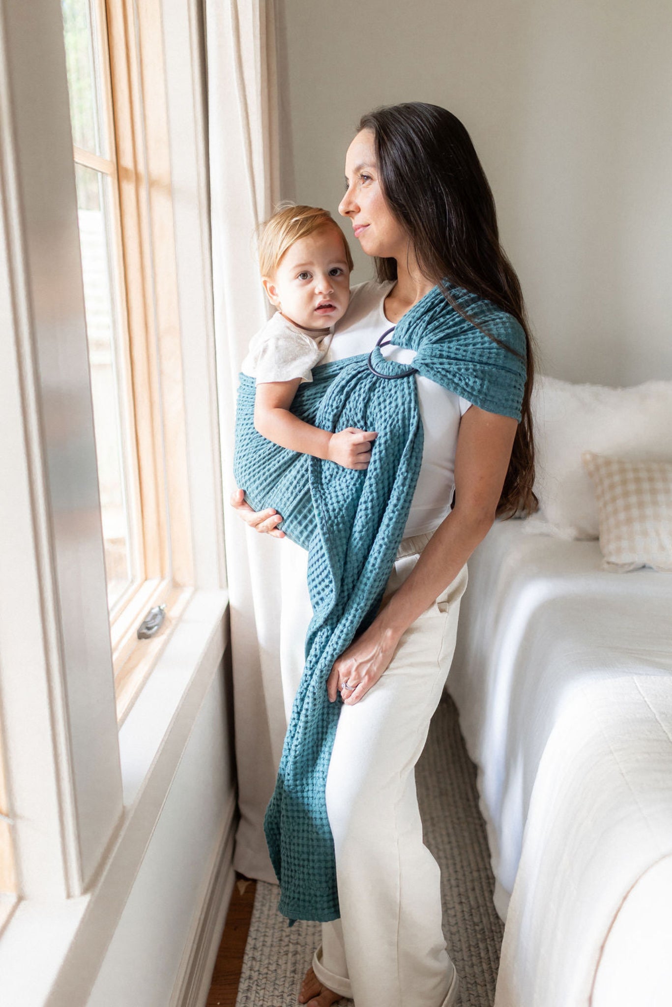 Woman holding a baby wrapped in a blue blanket ring sling baby carrier by a window