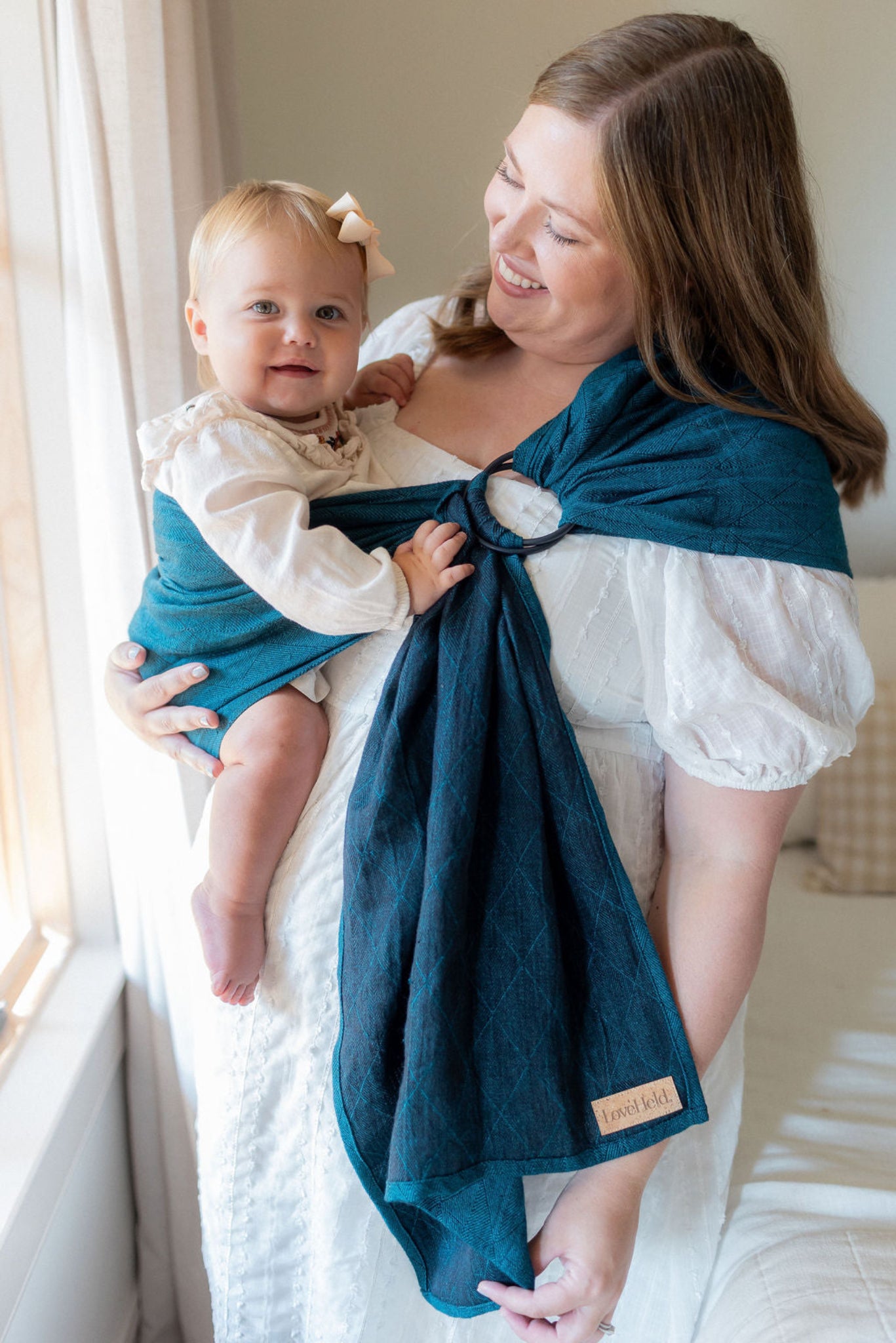 Woman holding a baby wrapped in a teal linen ring sling baby carrier in a bright room.