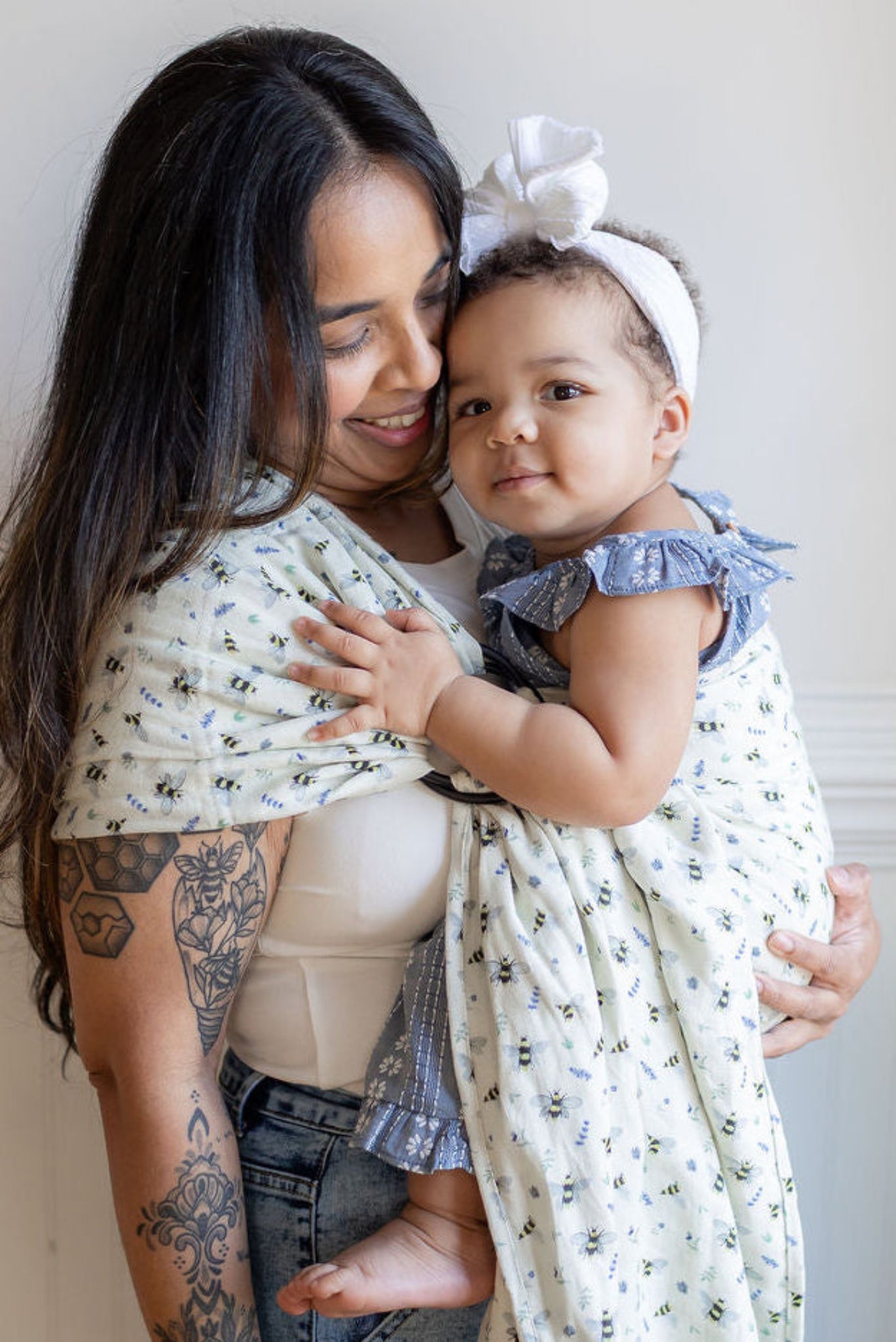 Woman holding a baby wrapped in a bee and floral wrap against a plain wall.
