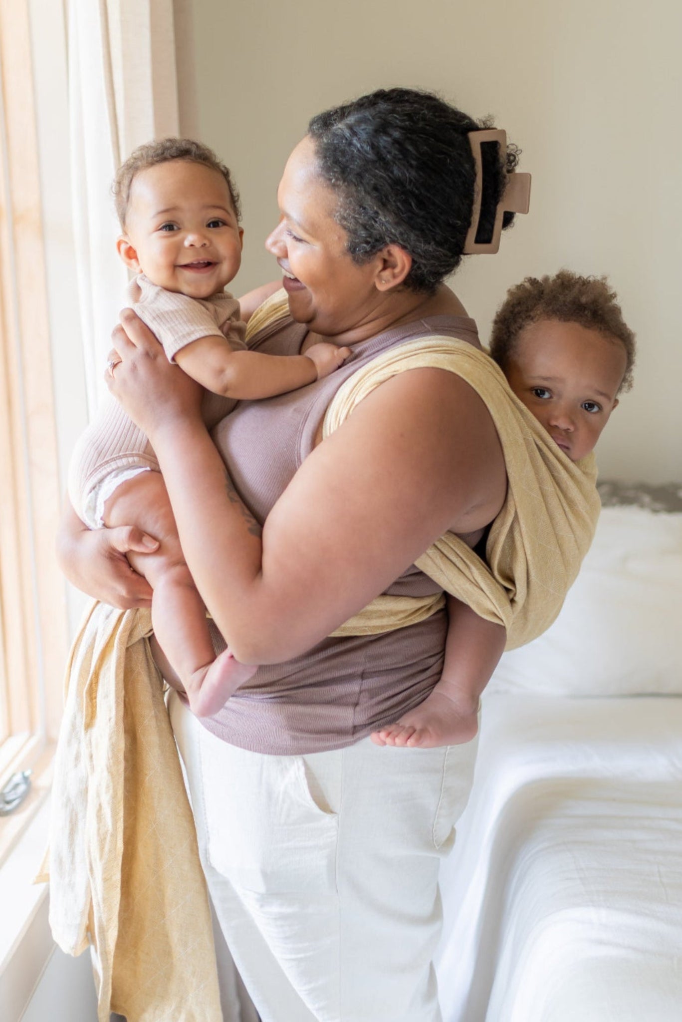 Woman holding two children in a room with a bed and window.