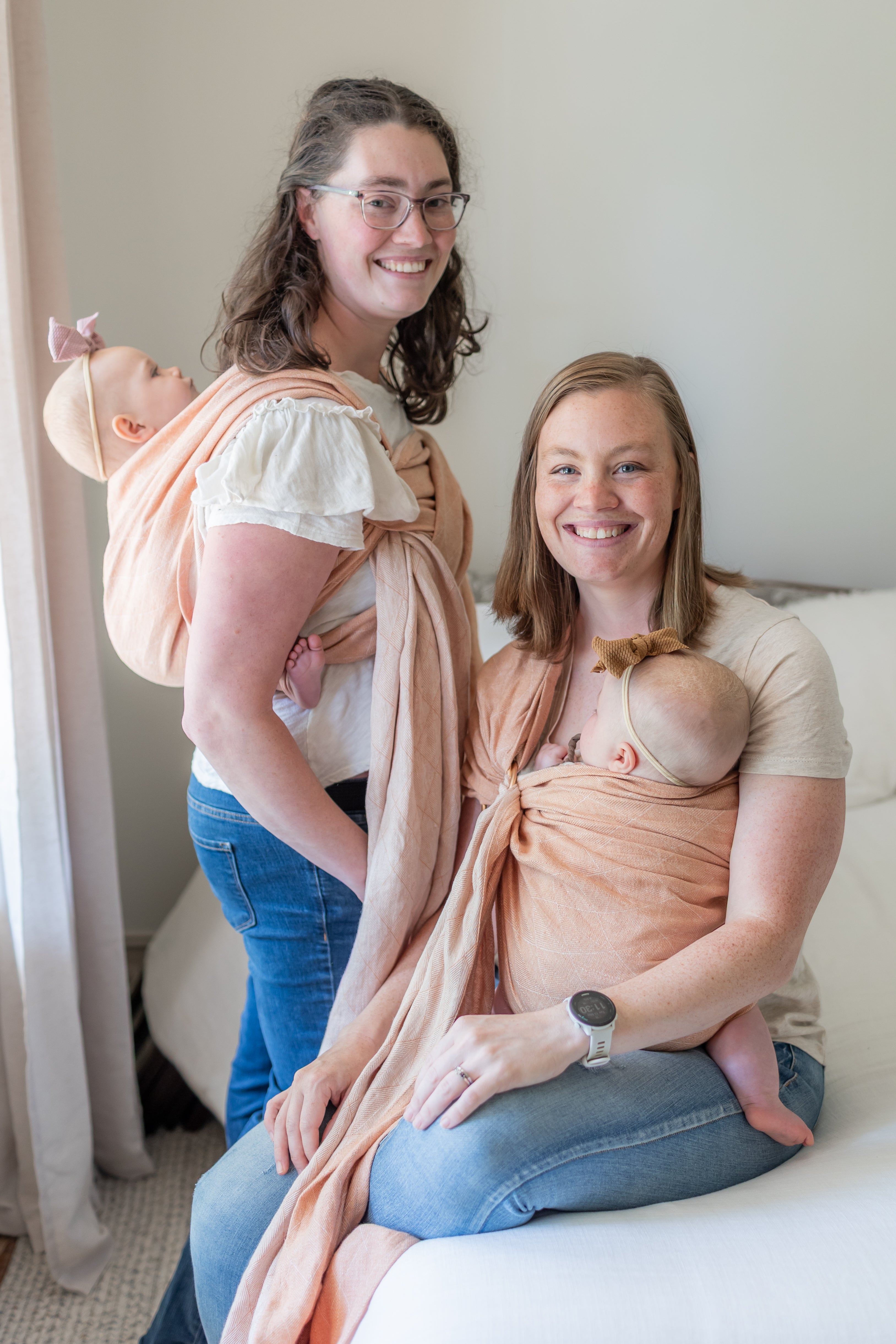 Two women with a baby in a sling and another baby being breastfed, smiling at the camera.